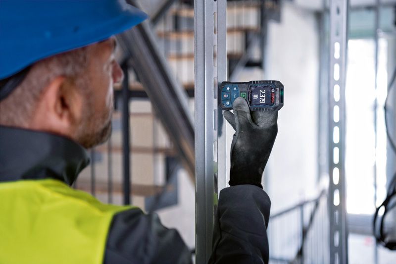 A person wearing safety equipment uses a laser measure on a metal beam.