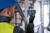A person wearing safety equipment uses a laser measure on a metal beam.