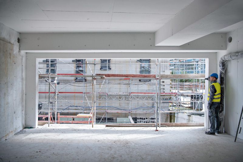 A person wearing safety equipment uses a laser measure to check distance inside a building shell.