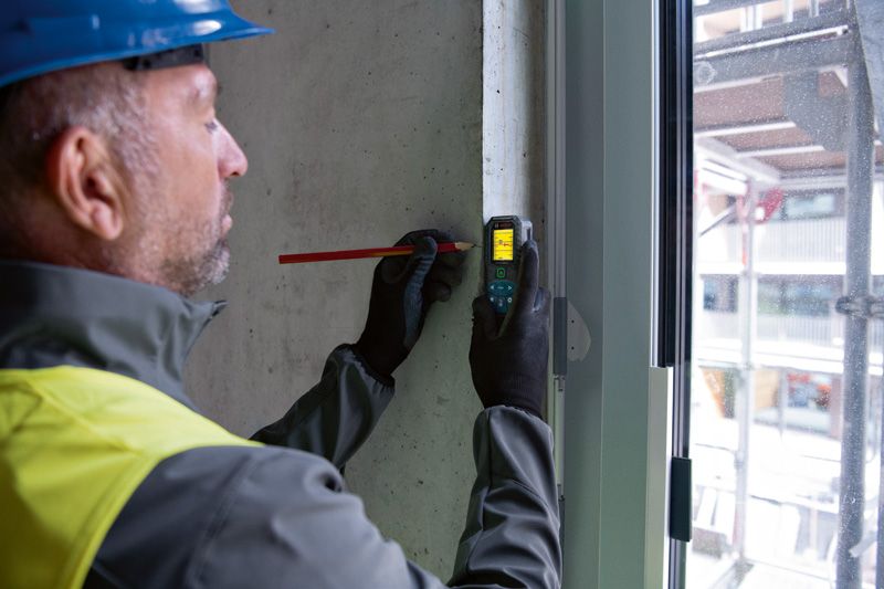 A person wearing safety equipment marks a wall using a laser measure.