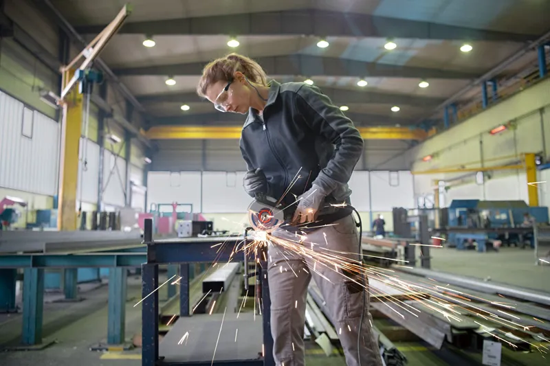 A person wearing safety equipment uses an angle grinder to cut metal in a factory.