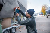 A person wearing safety equipment sands a boat hull outdoors with a random orbit sander.