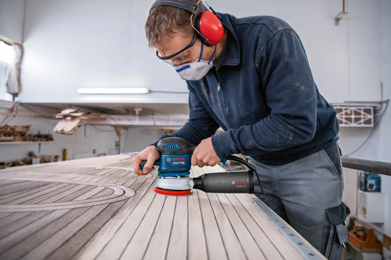 A person wearing safety equipment sands wooden planks with a random orbit sander.