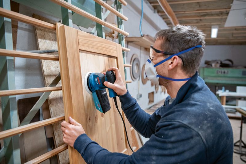Person wearing safety equipment sanding a wooden cabinet door with a random orbit sander.