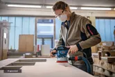 A person wearing safety equipment sands a wooden surface with a random orbit sander.