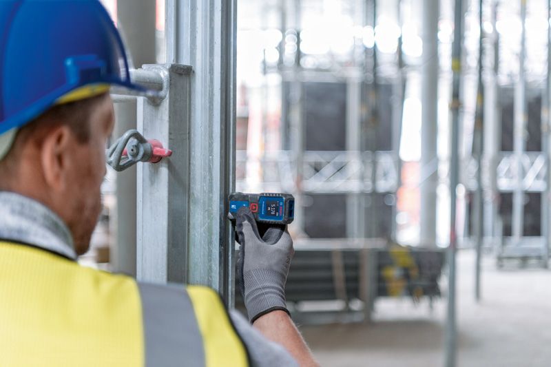 A person wearing safety equipment uses a laser measure against a metal beam.