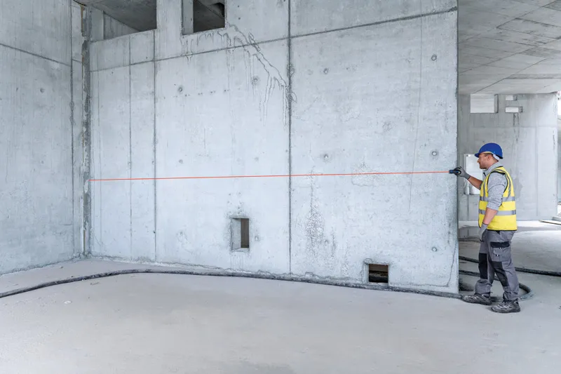 A person wearing safety equipment uses a laser measure to check distance on a concrete wall.