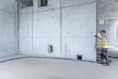 A person wearing safety equipment uses a laser measure to check distance on a concrete wall.