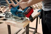 Person using a cordless circular saw to cut a wooden plank on a workbench.