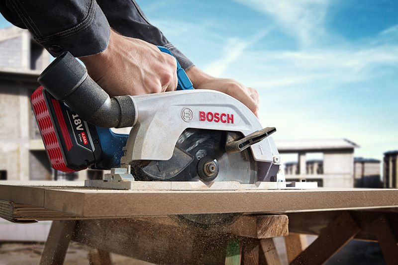 A person guides a cordless circular saw cutting through a wooden board at a construction site.