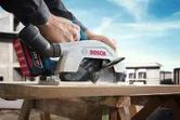 A person guides a cordless circular saw cutting through a wooden board at a construction site.