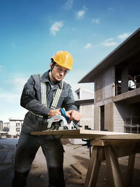 A person wearing safety equipment cuts wood with a circular saw at a construction site.