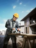 A person wearing safety equipment cuts wood with a circular saw at a construction site.