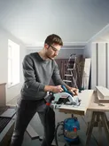 A person wearing safety equipment uses a circular saw to cut a wooden board.