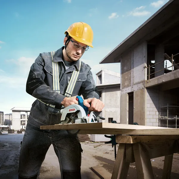 A worker wearing safety equipment uses a circular saw to cut wood at a construction site.