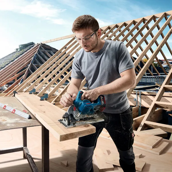 A person wearing safety equipment cuts a wooden board with a circular saw at a construction site.