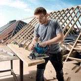 A person wearing safety equipment cuts a wooden board with a circular saw at a construction site.