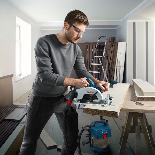 A person wearing safety equipment cuts a wood board with a circular saw on a workbench.