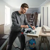 A person wearing safety equipment cuts a wood board with a circular saw on a workbench.