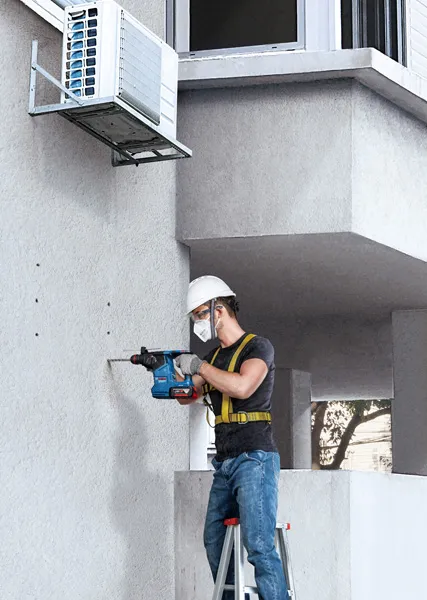 A person wearing safety equipment drills into a concrete wall beside an air conditioning unit.