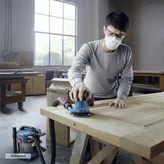A person wearing safety equipment sands a wooden door with a connected orbital sander.