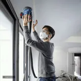 Person wearing safety equipment sands a ceiling near a window with a power sander.
