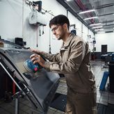 A person wearing safety equipment sands a car door with a power sander.