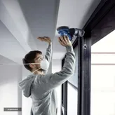A person wearing safety equipment sands a ceiling edge using a power sander.