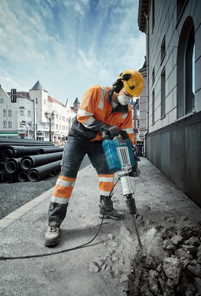 A person wearing safety equipment breaks pavement with a power breaker on a city street.