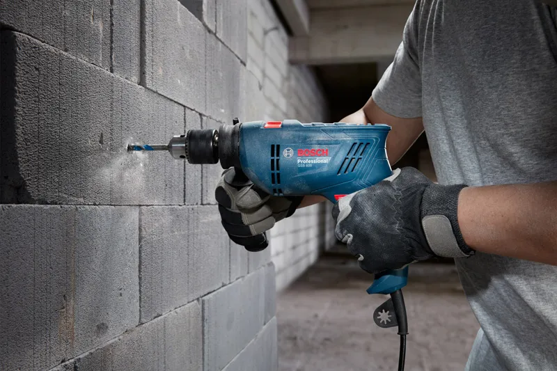 A person wearing safety equipment drills into a concrete block wall with an impact drill.