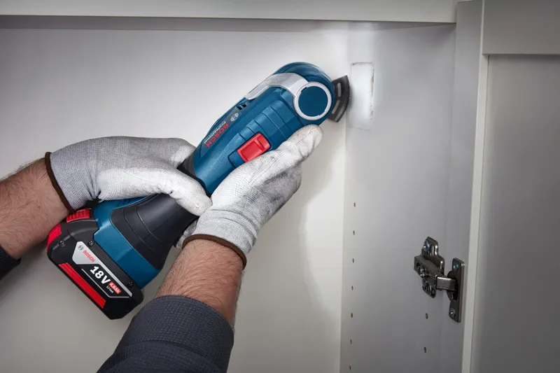A person wearing safety equipment uses a cordless power tool to cut a cabinet wall.