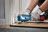 A person wearing safety equipment trims wood flooring with a cordless multi-tool.