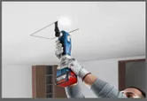 A person wearing safety equipment cuts a ceiling panel with a cordless oscillating tool.