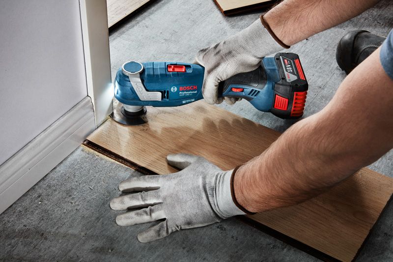 Person wearing safety equipment uses a multi-tool to cut flooring along a wall.