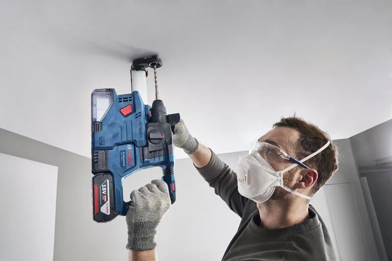 A person wearing safety equipment drills into a ceiling using a cordless rotary hammer.