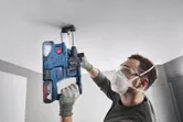 A person wearing safety equipment drills into a ceiling using a cordless rotary hammer.