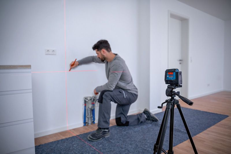 A person kneeling marks a wall aligned by a laser leveling tool on a tripod.