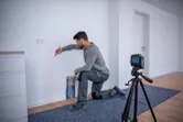 A person kneeling marks a wall aligned by a laser leveling tool on a tripod.