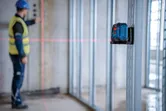 A person wearing safety equipment aligns wall studs using a laser leveling tool.