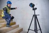 A person wearing safety equipment aligns a wall mark using a laser leveling tool on a tripod.