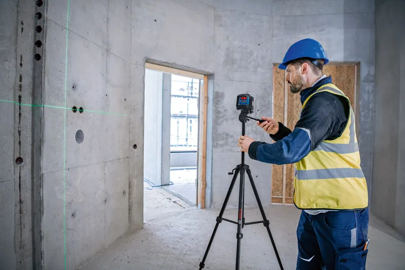 A person wearing safety equipment sets up a laser leveling tool on a tripod in a concrete room.
