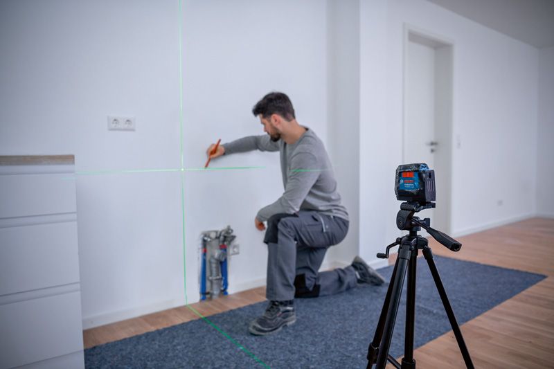 Person kneeling and marking a wall guided by a laser leveling tool on a tripod.