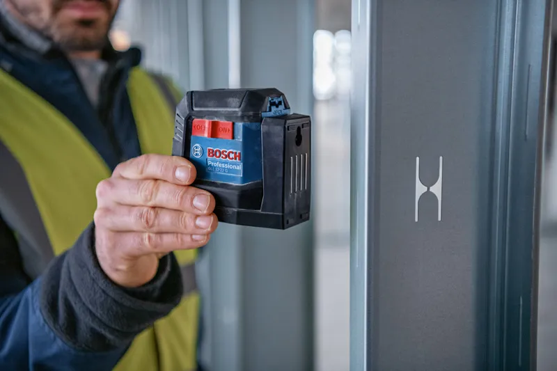 A person wearing safety equipment uses a laser leveling tool near a metal beam.
