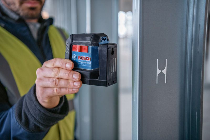 A person wearing safety equipment uses a laser leveling tool next to a metal beam.