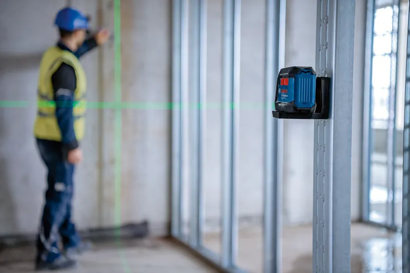 A person wearing safety equipment aligns framing studs using a green laser leveling tool.