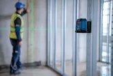 A person wearing safety equipment aligns framing studs using a green laser leveling tool.