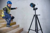 A person wearing safety equipment aligns a wall mark using a laser leveling tool on a tripod.