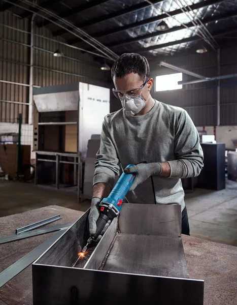 A person wearing safety equipment grinds metal inside an industrial workshop.