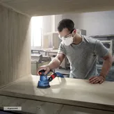 A person wearing safety equipment sands wood with a random orbit sander inside a workshop.