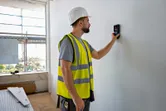 A person wearing safety equipment scans a wall with a detector tool.